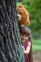 A sweet, pretty girl of 6 years old with long hair laughs cheerfully and looks out from behind a large tree. A large toy teddy bear peeks above her