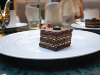 A piece of French opéra pastry in  white plate with coffee pot in background. It is made with layers of almond sponge cake soaked in coffee syrup, layered with ganache and coffee French buttercream