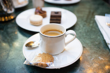 A cup of black espresso coffee with spoon and cookie, a plate of assorted dessert such as opéra pastry, macaron and mini tart in background on white table with spoon. - Powered by Adobe