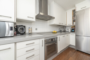 Furnished kitchen on one of the walls with white furniture, integrated stainless steel appliances, electric oven, gray countertop and dark flooring