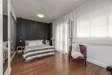 Fototapeta na wymiar Double bedroom en suite, chest of drawers with white drawers, TV attached to the wall, balcony with aluminum and glass door, armchair upholstered in violet silk, wooden floors