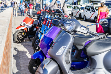 Several motorcycles and mopeds of all kinds and a row of blue electric scooters standing on a sidewalk in a European city