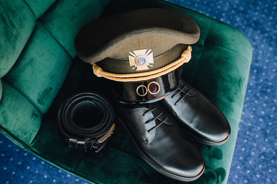 Gold rings, a cap, leather shoes, a belt, the uniform of a Ukrainian military groom on a chair. Wedding photography, accessories, details.