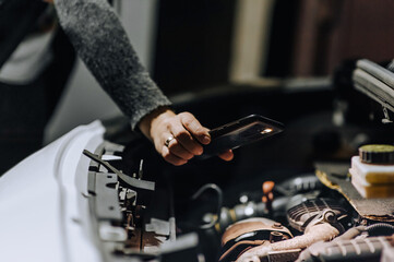 A woman mechanic repairs a car with a breakdown in the engine under the hood using tools at night, highlighting it with a flashlight on her phone or smartphone. Photography, portrait.
