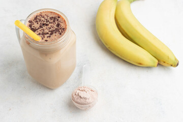 Glass jar of protein chocolate milkshake drink or smoothie and whey protein powder in measuring spoon, bananas on a white background. sport nutrition, bodybuilding food supplements
