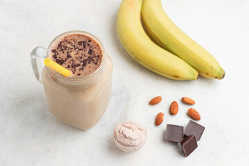 Glass jar of protein milkshake drink or smoothie and whey protein powder in measuring spoon, bananas, chocolate cubes, almond nuts on white background. sport nutrition, bodybuilding food supplements