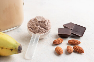 Chocolate whey protein powder in measuring spoon, glass jar of protein milkshake drink or smoothie, bananas, chocolate cubes and almonds on white background. sport nutrition, food supplements