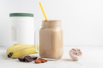 Glass jar of protein milkshake drink or smoothie and whey protein powder in measuring spoon, bananas, chocolate cubes, almond nuts on white background. sport nutrition, bodybuilding food supplements