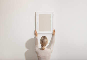 Blond young woman hanging a blank white wooden frame on the white wall. Minimalistic lifestyle,...