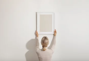 Blond young woman hanging a blank white wooden frame on the white wall. Minimalistic lifestyle, copy space for your text.. © Nika Votinova
