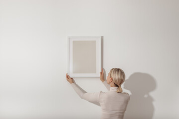 Blond young woman hanging a blank white wooden frame on the white wall. Minimalistic lifestyle, copy space for your text.. - obrazy, fototapety, plakaty