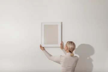Blond young woman hanging a blank white wooden frame on the white wall. Minimalistic lifestyle, copy space for your text. © Nika Votinova