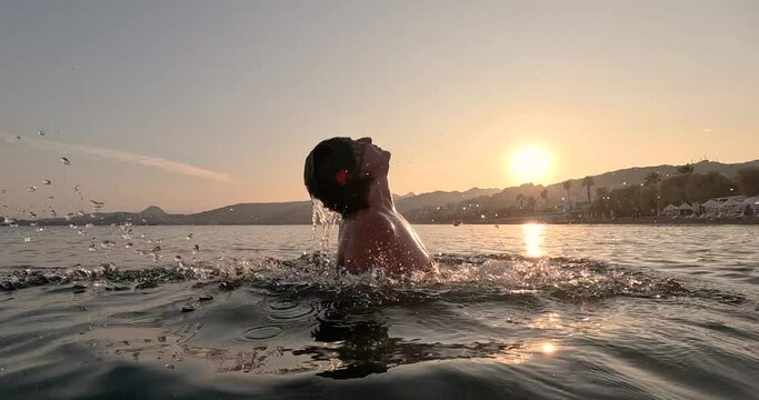 Beautiful woman throwing her hair back with water in the ocean at sunset. Slow motion video in 4K resolution. Side view. Summer holiday concept.