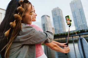 Two girls making video with smartphone and selfie stick while standing on city waterfront