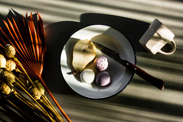 Still life in dark key with fruit, sweets, cup on table top view. Breakfast in a morning with contrast shadows. Rustic kitchen. A ripe pear cut in half, a knife, lilac violet macaroons. French desert.