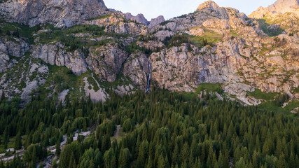 A high waterfall cascades in a mountain gorge. Coniferous trees, Christmas trees and grass grow. The river runs. Hiking trails. Top view from a drone. Sunset. Burkhan Bulak Waterfall, Kazakhstan © SergeyPanikhin