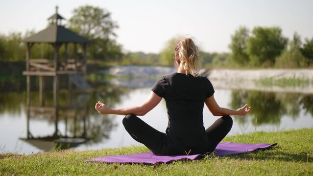 Back view of a woman sitting in the lotus position and meditating by a lake in nature