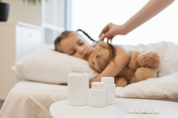 Various jars of pills standing on white bedside table with sick caucasian girl on blurred background. Tired kid lying on comfy bed with toy bear and measuring temperature with thermometer.