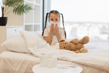 Sad ill girl in pyjama sitting on soft bed with teddy bear toy under blanket and blowing nose into handkerchief. Focus on white coffee table full of various medication and electronic thermometer.