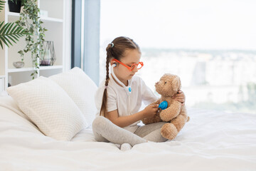 Cute girl dressed in casual clothes sitting on comfort bed and listening heartbeat of fluffy teddy bear with toy stethoscope. Pretty kid imagining herself as doctor and treating her beloved patient.