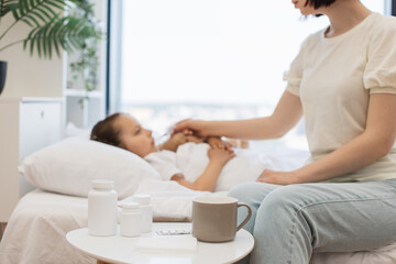 Fototapeta premium Focus on bedside table with cup of hot beverage, paper tissues and medical pills standing near white bed. Blurred background of caring mother measuring daughter's fever with digital thermometer.