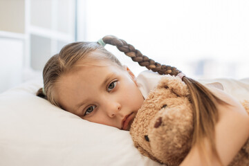 Caucasian little girl with gloomy look sleeping in white bed and seeking solace in embrace of her teddy bear. Sick kid with two braids suffering from cold and flu symptoms at home.