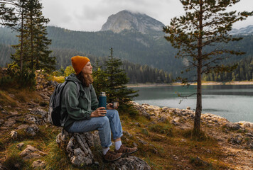 Young cheerful woman traveler with a backpack and thermos on the shore of a lake in the mountains....