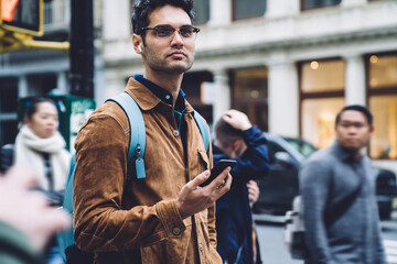 Thoughtful Hispanic young man standing on busy sidewalk with smartphone