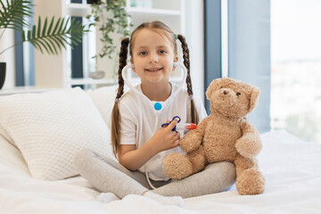 Smiling girl using toy syringe for injecting fluffy teddy bear with plastic stethoscope in ears on comfort white bed. Beautiful caucasian child imagining herself as doctor and performing procedures.