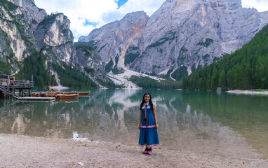 happy Asian woman tourist standing on the coast of Braies lake. Dolomites, Italy. Landscape of famous lake with beautiful reflection in water, trees, sky with clouds. wooden boat hut. Travel.