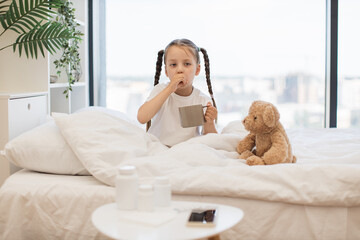 Adorable caucasian baby sitting in bed with teddy bear and drinking hot tea. Beautiful child having cold and treating while staying at home with attractive panoramic view of city.