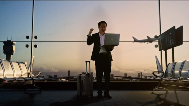 Full Body Of Asian Businessman With Rolling Suitcase Waits For Flight In Boarding Lounge, Looking At A Laptop And Celebrating, Airport Terminal With Airplane Takes Off Outside The Window
