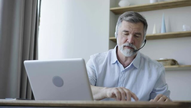 Mature businessman wear headphones making video conference business call on laptop speaking communicating by webcam, male teacher manager talking do online chat on computer sit at home office desk