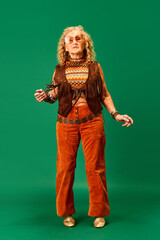 Hippie style. Extravagant senior woman in stylish cloths and accessories standing over green studio background. Concept of human emotions, fashion, elderly people, lifestyle, creativity. Ad
