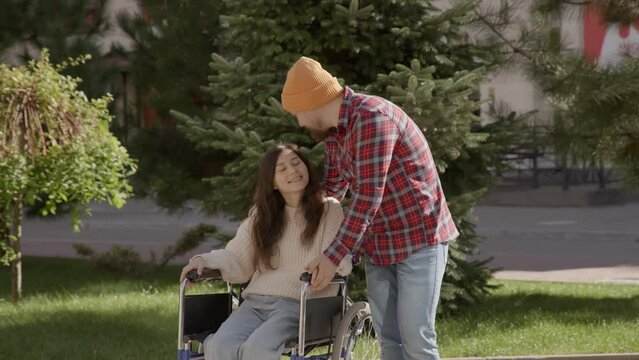 Young smiling man accompany pretty woman in a wheelchair portrait. Couple in love. Invalid girl have a nice day dating with her boyfriend walking outside in park. Concept of togetherness, support