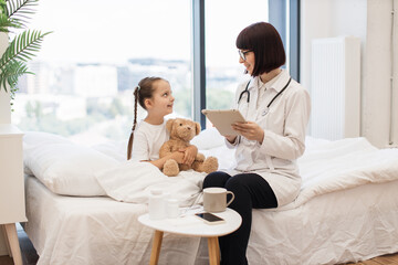 Competent female pediatrician prescribing medicine on modern tablet for sick child in spacious bedroom. Smiling girl holding teddy bear and preparing for intensive treatment at home.