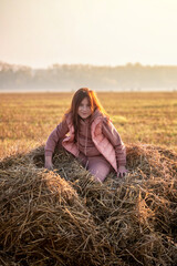 Naklejka na ściany i meble Cute teenage girl in field with wheat clippings and haystacks. morning, fog