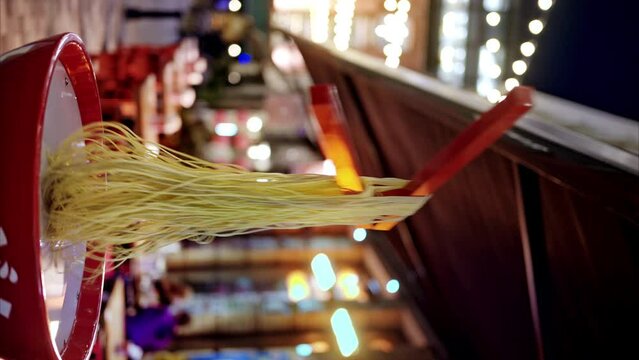Vertical view of moving Chinese chopsticks with noodles in the Chinatown at evening in London, United Kingdom. Pedestrian street with decorations, illumination and moving people on the background