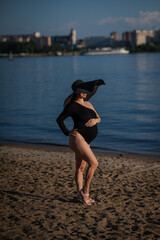 Pregnant woman in a large straw hat and black swimsuit posing on the beach.