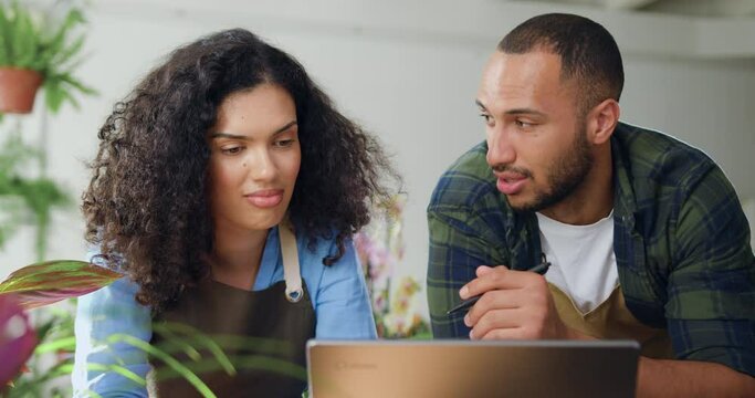 Close Up. African American Young Couple Florists In Aprons Working In Flower Store And Talking Typing On Laptop. Woman Working At Laptop Together With Man Florist Using An Online Service To Accepting