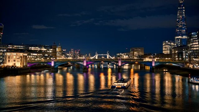 View of Thames River at night in London, United Kingdom. Floating boat, bridges and Tower Bridge in the distance, Shard skyscraper and other buildings on the both sides, a lot of illumination