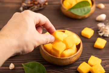 Dried mango cubes in wooden bowls on brown wooden with hand, side view, selective focus.
