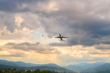 Fototapeta na wymiar White passenger plane takes off against a beautiful sky with golden clouds and green mountains. Travel by air