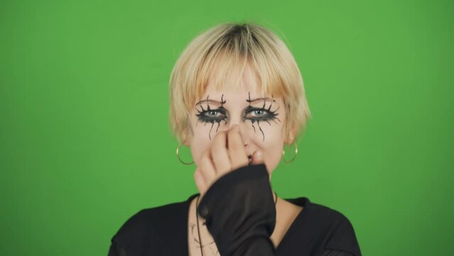 Portrait of a goth girl in black clothes and special makeup on a green background. She looks at the camera, sticks out her tongue. Chroma key.