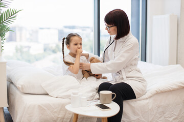 Obraz na płótnie Canvas Experienced nurse sitting on bed and using stethoscope while small lady covering mouth with hand and coughing in bedroom. Concept of home treatment and friendly relations between kid and doctor.