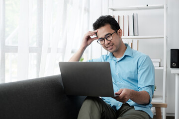 Young attractive man sitting on sofa at home working on laptop online, using internet, smiling, happy mood, freelancer, free leisure time, relaxed, modern job lifestyle