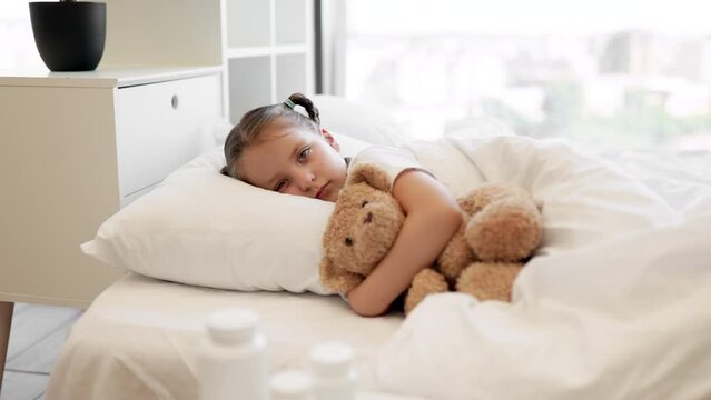 Tired caucasian little girl lying in soft bed with teddy bear in embrace and feeling sick. Coffee table with pills and thermometer standing near. Health care and disease concept.