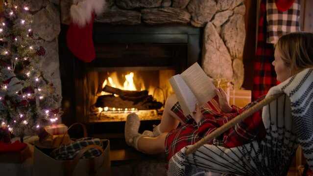 Relaxed female sitting in chair and enjoying her winter holiday weekend in cabin 4K. Cinematic shot of young woman reading book and relaxing at cozy burning fireplace at home decorated for Christmas