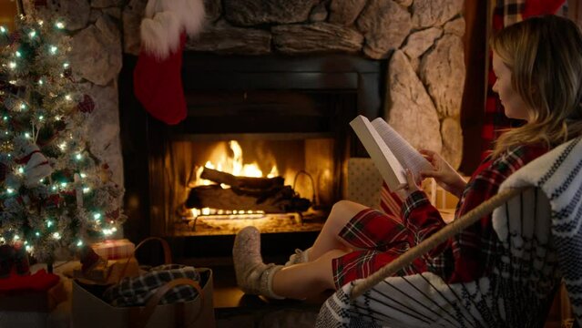 Cozy burning fireplace at home decorated for Christmas. Relaxed female sitting in chair and enjoying her winter holiday weekend in cabin 4K. Cinematic shot of young woman reading book and relaxing