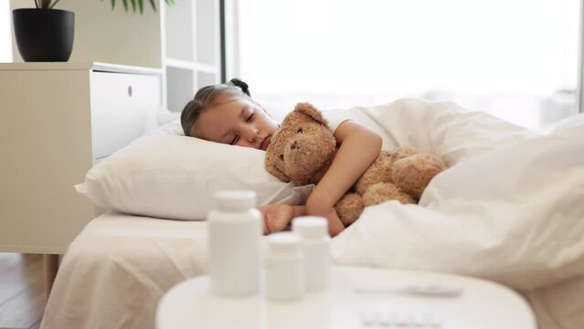 Cute caucasian girl sleeping sweet on white soft pillows under blanket and hugging her teddy bear friend. Pretty kid with two braids regaining energy during disease at home.